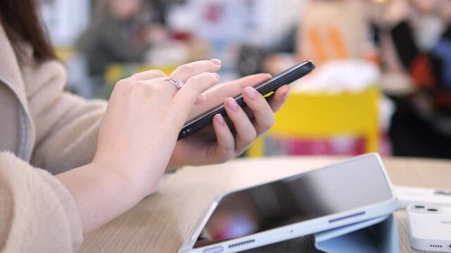 Person uses smartphone while seated at table with other devices. Fingers tap screen with focus and intent. Background blurred, suggesting casual public space. Suitable for tech, communication