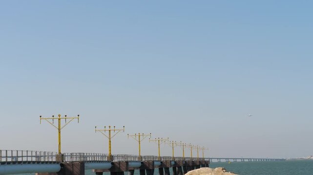 Runway approach lighting pier extending over the sea with a far airplane descending for landing. Sunny day, clear blue sky. Static wide shot of Hong Kong airport ALS