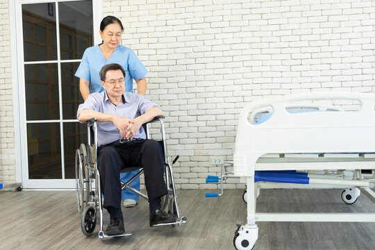 Smiling nurse assisting senior man to get up from bed. Caring nurse supporting patient while getting up from bed and move toward wheelchair at hospital. Helping elderly disabled asian man standing up.