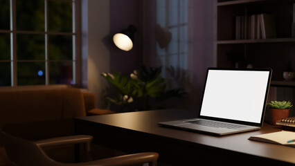 Dark home office with white screen laptop on wooden table beside bookshelf illuminated by floor lamp © bongkarn