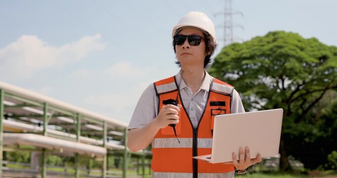 Asian adult female technician speaks through walkietalkie while holding laptop at oil refinery, managing emergency communication, pipeline safety inspection, compliance monitoring workflow report