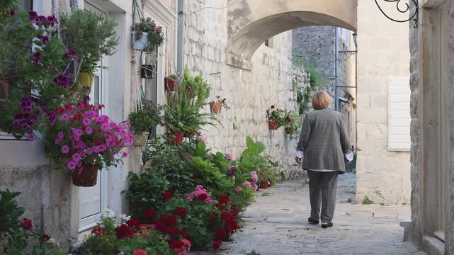 Mature female tourist seen from behind walking through a narrow stone street with flowers in a historic Mediterranean old town. Europe travel and sightseeing scene.