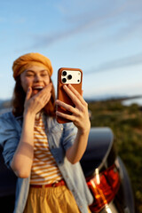 Obraz premium Smiling woman outdoors taking a selfie with a smartphone, wearing a striped shirt and orange beanie. Casual lifestyle moment captured during golden hour, radiating joy, travel, and wellness.