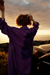 Obraz premium A woman wearing a purple robe stands with her back to the camera beside a car at sunset, raising her arms in a moment of reflection, freedom, and self care.