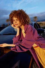 Obraz premium A stylish woman leans on a sleek car at dusk, wearing a purple jacket and casual outfit. She smiles softly, capturing a relaxed, confident lifestyle moment in the evening light.