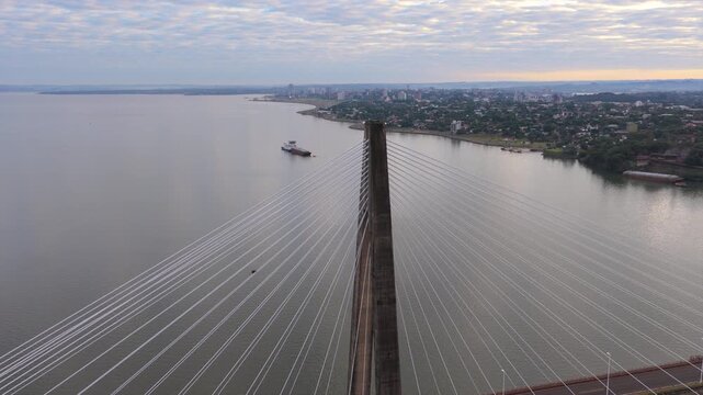 Drone footage of a cargo ship passing beneath the San Roque Gonzalez International Bridge with Encarnacion city in the background.