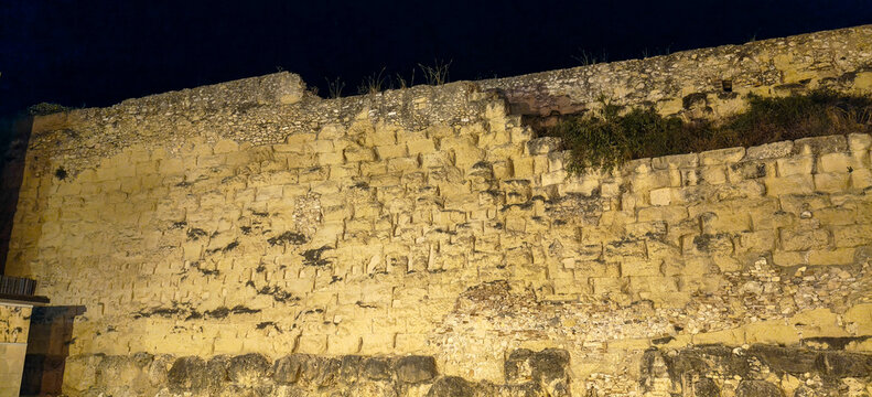 Muralla romana hist&oacute;rica iluminada de noche junto a una torre de piedra, con jardines p&uacute;blicos y edificios urbanos al fondo en la imagen