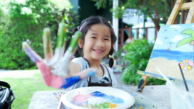 Cute happy Asian little girl showing her colorful painted hand, smiling child enjoying creative art workshop and beach canvas painting activity in the park