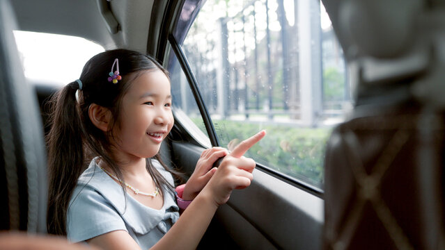 Happy Asian little girl looking out of the car window during a family road trip, smiling child enjoying summer vacation travel and transportation concept.