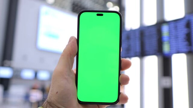 Hand holds phone with green screen at airport. Background shows flight information boards. Focus on device for overlays or app demos. Ideal for travel, tech, or digital marketing projects