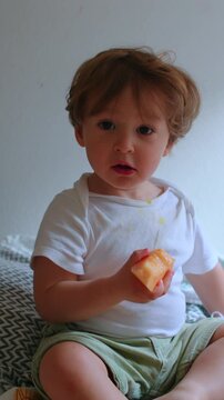 Toddler eating fresh melon while relaxing on bed at home, highlighting healthy snacking, childhood curiosity, comfort and everyday family life