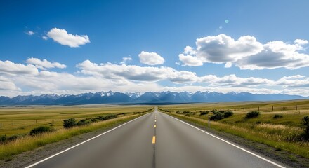Fototapeta premium Endless open road leading towards majestic snow capped mountains under a bright blue sky with fluffy clouds.