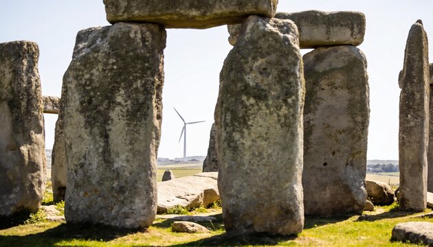 Framing large worn upright stones revealing distant wind turbine on rural plain, lintel, lichen
