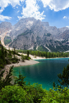 Turquoise water of Lake Braies with gravel shore surrounded by green pine forests and towering rocky mountain cliffs under a cloudy blue sky, scenic Pragser Wildsee landscape