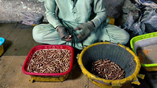 Unrecognizable worker sitting on the floor of a bullet factory, meticulously counting and sorting ammunition by hand into baskets in a poor, developing country manufacturing facility