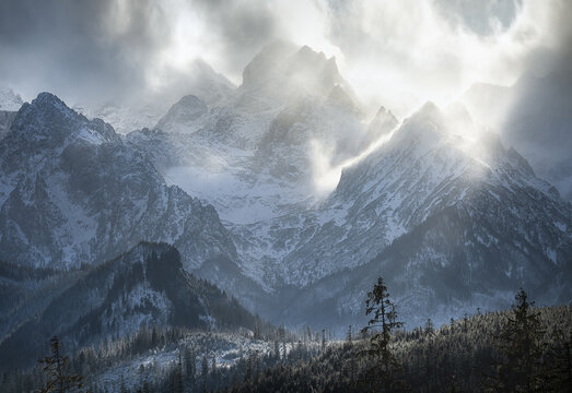 View of the snow-covered peaks of Tatra National Park with dramatic clouds and sunlight rays over the evergreen forest in Zakopane, Wojewodztwo Malopolskie, Poland.
