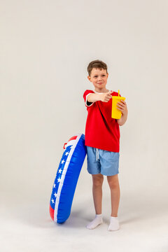 Little boy wearing a red t-shirt and blue shorts stands with an inflatable pool ring, holding a yellow drink, smiling cheerfully on white background. 4th of July pool party in USA