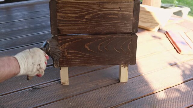Brown oil for protection of wood by staining. Painting fence for protecting and pests. Woodworker applying finish to pine board box. Repairman varnishing with brush wooden fencing wall plank surface.