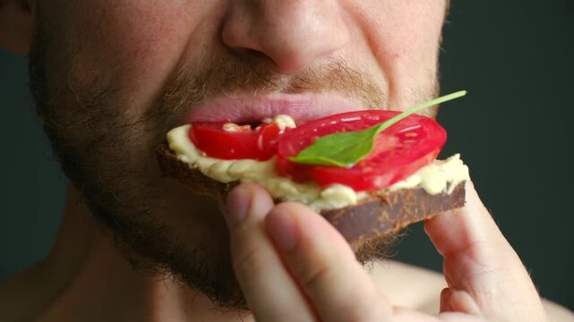 Extreme close up bearded man mouth eating healthy vegan bruschetta with hummus fresh tomato spinach on rye bread, showing process biting, chewing the sandwich. Man eating healthy vegan tomato sandwich