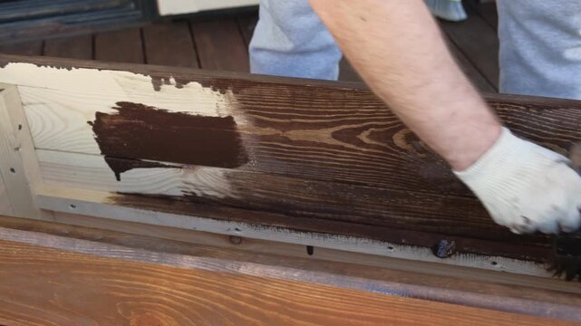 Brown oil for protection of wood by staining. Painting fence for protecting and pests. Woodworker applying finish to pine board box. Repairman varnishing with brush wooden fencing wall plank surface.