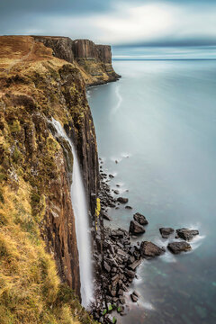 View of Kilt Rock Waterfall cascading down steep basalt cliffs into the sea under an overcast sky in Portree, Highland Council, United Kingdom.