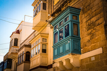 Obraz premium looking up at mdina Maltese homes on narrow alleyway with balcony and windows with green painted wood and limestone bricks on sunny summer day with clear blue skies overhead