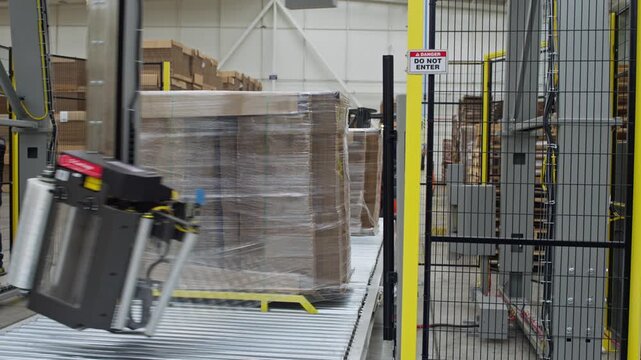 Wrapped pallet of cardboard sheets moving through automated warehouse conveyor system, plastic shet rotates around with mechanized arm