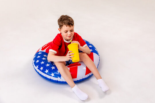 Little boy in red shirt sits on a round inflatable pool float featuring an American flag print design, holds yellow drink cup, smiling, white background, copy space. 4th of July pool party in USA