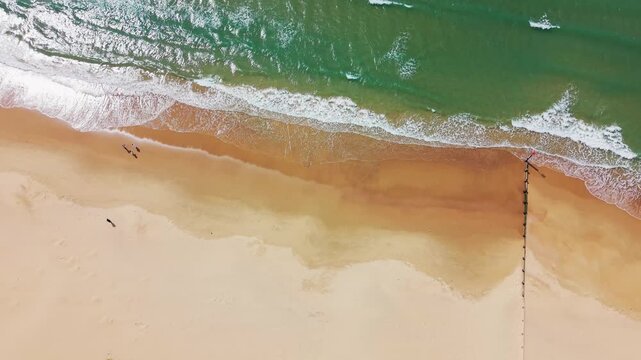 Top down aerial drone footage of Bournemouth beach showing waves rolling onto a sandy shoreline, turquoise sea, white surf, beach walkers and a coastal groyne. Useful for themes of UK beaches