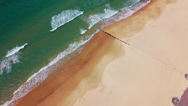 Top down aerial drone footage of Bournemouth beach showing turquoise sea, white waves, sandy shoreline patterns and a coastal groyne stretching across the beach. Useful for themes of UK beaches
