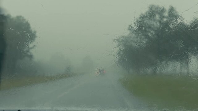 Close-up POV inside a car during heavy downpour with windshield wipers sweeping across the glass as cars approach with headlights on in a dark, grey, moody stormy environment.