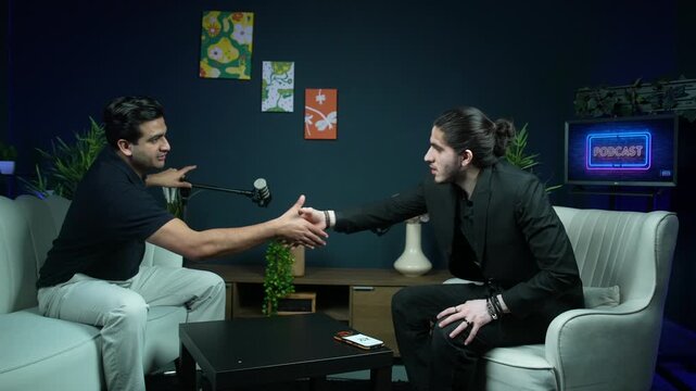 Two men greet with a handshake in a modern podcast studio, seated across table