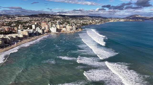 Aerial coastal city shoreline waves crashing, drone pans along turquoise ocean edge beside highrise