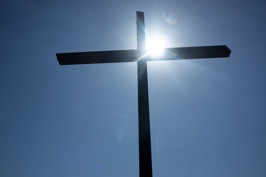 Croce scura in silhouette illuminata dal sole che irradia raggi di luce, fotografata contro un cielo limpido in una scena simbolica che evoca fede, speranza e profonda spiritualit&agrave;