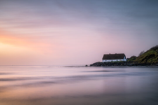 View of a traditional white thatched cottage on the rocky shore of Laytown Beach during a soft pastel sunrise with calm water Laytown, County Meath, Ireland.