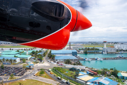 Seaplane propeller spins beside a red engine over Male harbor and marina near Velana International Airport, with TMA docks, white terminals, runway, boats, and roads visible.