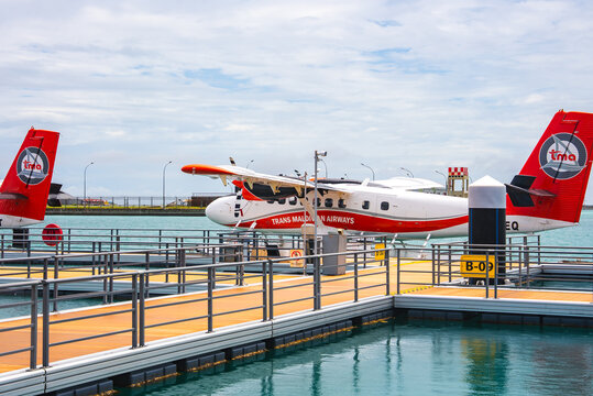 Trans Maldivian Airways seaplane docked at pier B 09 near Velana International Airport, Male, Maldives. Pontoons rest on a turquoise lagoon under a bright, slightly overcast sky.