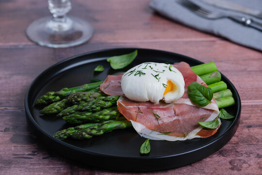 A brunch dish features a poached egg placed on slices of ham and fresh asparagus. The plate has green basil leaves as a garnish. Healthy eating concepts. side view