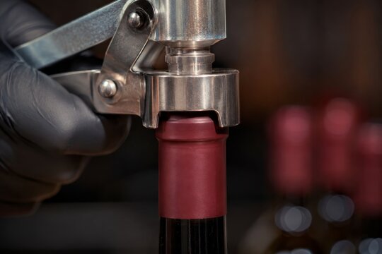 Winemaker using metal capper machine to seal wine bottle with foil cap. Process of bottling premium red wine in winery. Professional production and food industry equipment concept.