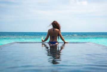 Fototapeta premium A woman with long hair relaxes at an infinity pool in the Maldives, holding a drink as soft daylight frames a centered scene of turquoise sea and pale sky.