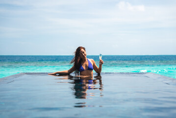 Obraz premium A woman relaxes in an infinity pool holding champagne at a Maldives resort, with turquoise ocean and gentle waves beyond the edge under bright daylight.