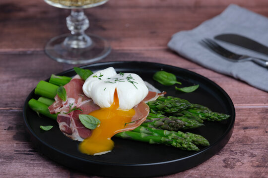 A dish features a poached egg on top of slices of ham and asparagus on a wooden table. A glass of drink is seen in the background. Healthy eating concepts. side view