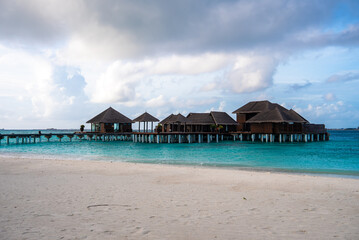 Fototapeta premium Thatched roof water villas on stilts connect by a long wooden boardwalk in the Maldives. Powdery white sand, calm seas, and cloud dappled skies frame the open horizon.