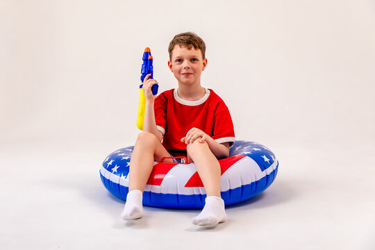 Little child boy wearing a red shirt and white socks sits on a large inflatable pool float featuring an American flag print design, holding toy water gun on white background. 4th of July party in USA