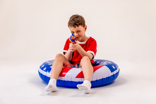 Little child boy in red shirt sits on a large inflatable pool float featuring an American flag print design, aiming a toy water gun at camera on white background. 4th of July party in USA