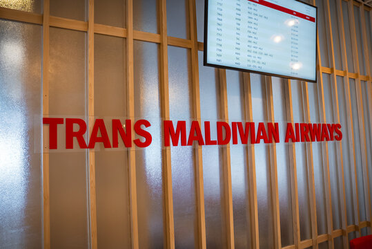 Trans Maldivian Airways lettering and a departures board appear in Male, likely at Velana International Airport. Soft even light meets wood slats and frosted panels.