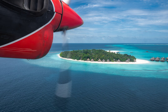 Aerial view from a red seaplane engine shows a Maldives atoll with white sand, turquoise lagoons, overwater villas, palm trees, and deep ocean under bright midday light.
