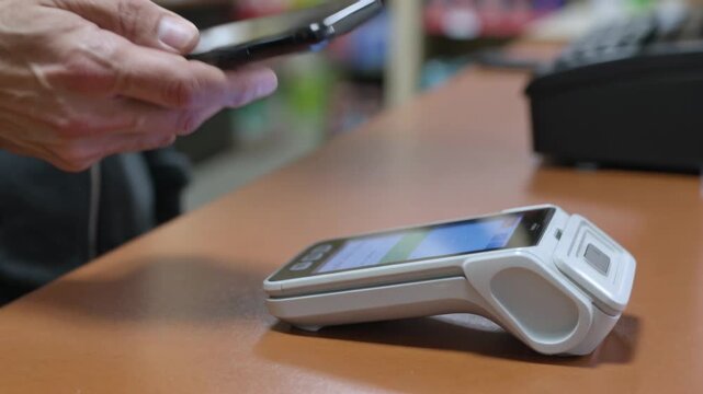 Starting NFC payment, man lowering phone over shop counter holding near white reader in dark hoodie