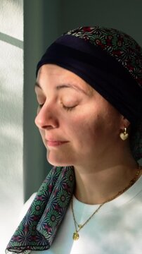 Woman in headscarf gazing through window during recovery period, reflective lifestyle portrait expressing resilience, hope, determination and personal healing