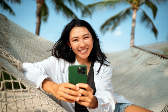 Happy young woman talking on her smartphone at a tropical beach, enjoying vacation, remote work